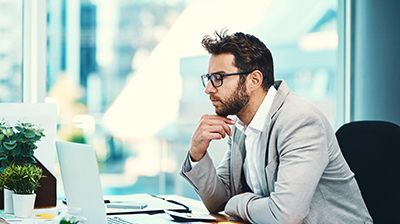 Man in business attire sitting at desk looking at laptop while resting chin on hand.