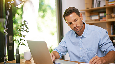 Man in business casual attire sting at desk with laptop and writing on papers.