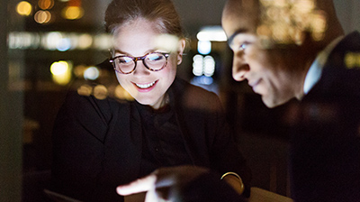 Man and woman in business attire sitting next to a window at night discussing over a laptop.