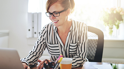 Woman in business casual attire sitting at a desk and smiling while using a laptop.