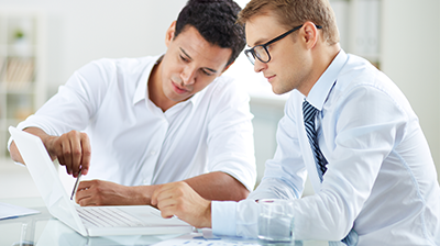 Two men in business attire at a desk looking over a laptop; One points to screen while the other looks on