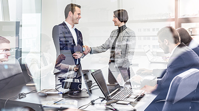 A business man and woman shaking hands in the reflection of a conference room where business people are having a meeting
