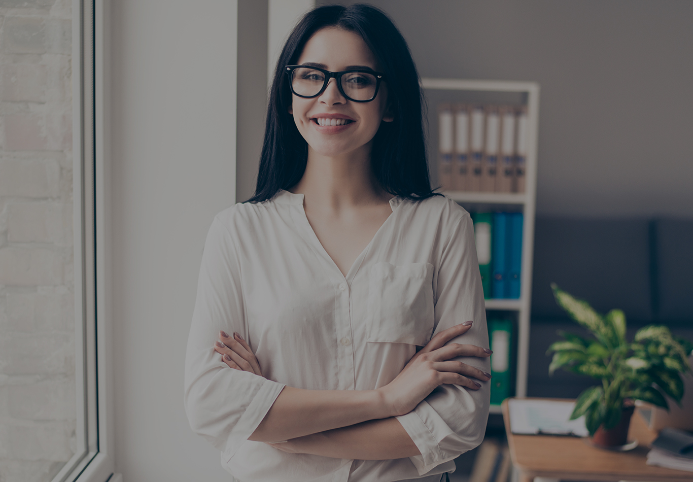 Woman in casual attire  in an office wearing large glasses with arms folded over her chest, she is smiling at the camera. 